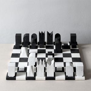 Op de afbeelding: Een modern schaakspel met zwarte en witte stukken op een schaakbord. De stukken zijn geometrische vormen in zwart en wit, wat een minimalistische esthetiek creëert. Het bord heeft een klassiek zwart-wit dambordpatroon.