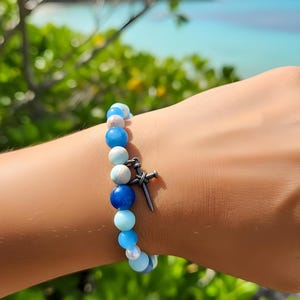 May include: A bracelet with blue, white, and pink beads, and a silver-coloured cross charm. The bracelet is worn on a wrist, with a blurred background of green foliage and the sea. The beads are of various sizes.