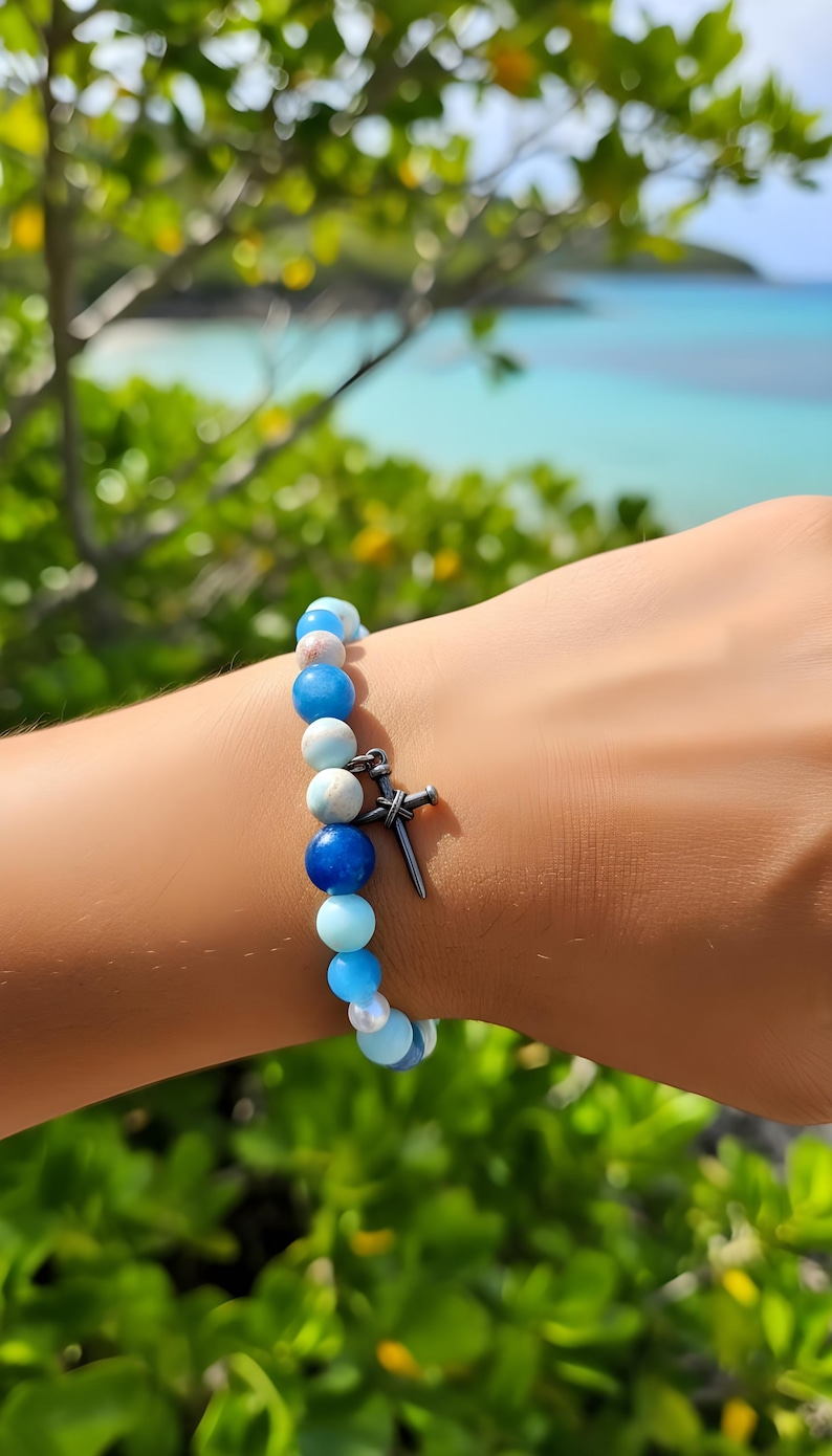 May include: A bracelet with blue, white, and pink beads, and a silver-coloured cross charm. The bracelet is worn on a wrist, with a blurred background of green foliage and the sea. The beads are of various sizes.