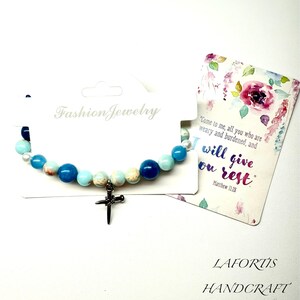 May include: A blue beaded bracelet with a silver cross charm. The bracelet has blue and white beads. A card with floral designs and the text "I will give you rest" is next to the bracelet. The words "Fashion Jewelry" are also visible.