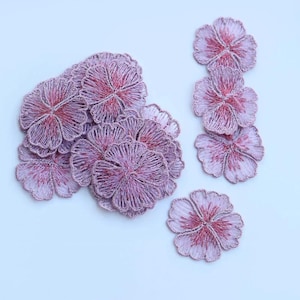 May include: A collection of embroidered flower patches in shades of lavender and pink. The floral designs feature detailed stitching, with a lighter lavender hue on the petals and a deeper pink in the center. These patches are ideal for fabric crafts and embellishments.