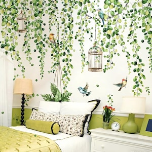 Puede incluir: Una escena de dormitorio con una pared blanca adornada con calcomanías de vid verdes, jaulas para pájaros y pegatinas de pájaros de colores. La cama tiene almohadas blancas, un cojín verde y una almohada estampada. Dos lámparas flanquean la cama.
