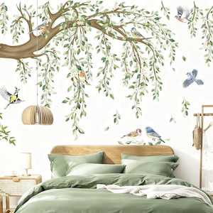 Op de afbeelding: Een slaapkamer met een muurschildering van een boom met groene bladeren en vogels. Het bed heeft groene beddengoed en kussens. Een houten hanglamp hangt aan het plafond. Een nachtkastje en een kledingrek zijn ook zichtbaar.