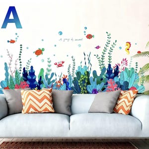 May include: A wall decal featuring an underwater scene with colorful seaweed, fish, a seahorse, and a turtle. The design includes the text "A group of seaweed" and a large blue letter "A". The decal is placed above a white couch with decorative pillows.