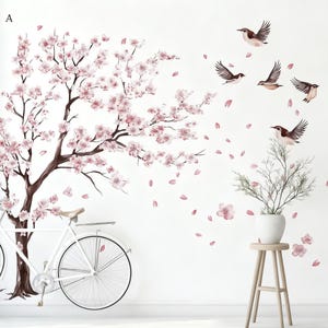 May include: Wall decal featuring a blossoming cherry tree with pink flowers and falling petals. Includes a white bicycle and several brown and white birds in flight. A small plant in a white pot sits on a wooden stool.