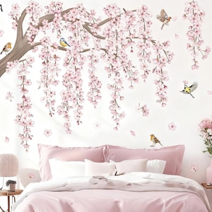 Op de afbeelding: Een muursticker met een kersenbloesemboom met roze bloemen en bruine takken. Verschillende kleine vogels zitten op de takken of vliegen rond de bloesems. De sticker is geplaatst boven een bed met roze beddengoed.