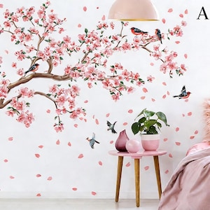 Op de afbeelding: Muursticker met een bloeiende kersenboom met roze bloemen en bruine takken. Verschillende vogels zitten op de takken. Roze bloemblaadjes en vogels zijn verspreid over de witte muur. Een kleine roze tafel heeft een plant en vazen.