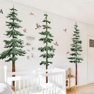 May include: Wall stickers depicting three green and brown pine trees with flying birds. The decals are on a white wall in a nursery, with a white cot and small wooden furniture in view.