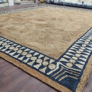 May include: Large rectangular jute rug with a natural tan color and a geometric border. The border features a repeating pattern of navy blue triangles and circles, with a tan background. The rug has a textured surface and a decorative design.