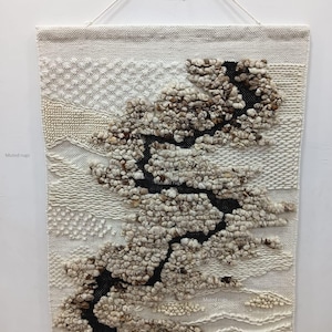 May include: A woven wall hanging with a textured design. The artwork features a black, vertical element with a tree-like shape, surrounded by beige and brown textured elements. The top of the hanging has the text "Muted rugs".