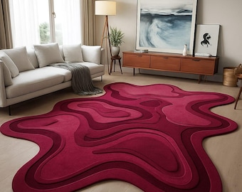 May include: A modern, abstract area rug in shades of red. The rug features a layered, organic design with varying depths and tones, creating a three-dimensional effect. It's placed on a light-colored wooden floor in a living room setting.