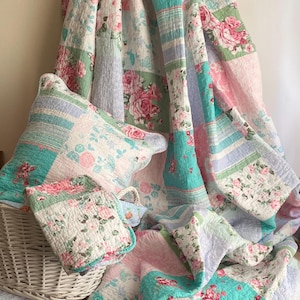 May include: A pastel-coloured patchwork quilt and cushion set. The quilt displays large squares with floral and striped patterns in pink, green, and blue. A small folded blanket and a white wicker basket are also visible.