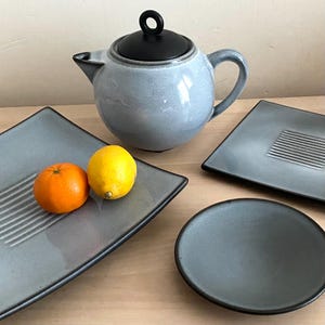 May include: A grey ceramic teapot with a black lid and handle is surrounded by grey square plates and a round plate. An orange and a yellow lemon sit on a square plate. The plates have a black trim.