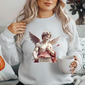 Puede incluir: Sudadera gris claro con un dise&ntilde;o de estatua de Cupido. La estatua tiene alas, arco y flecha, y un drapeado rosa. El letrero dice "Overbooked for Valentine's".