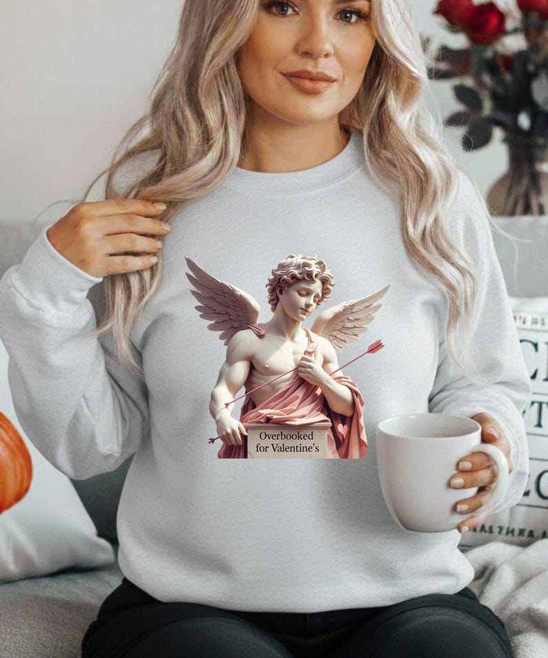 Puede incluir: Sudadera gris claro con un dise&ntilde;o de estatua de Cupido. La estatua tiene alas, arco y flecha, y un drapeado rosa. El letrero dice "Overbooked for Valentine's".