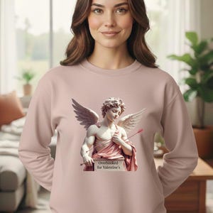 Puede incluir: Sudadera rosa claro con la imagen de un Cupido alado sosteniendo una flecha y un cartel con la frase "Overbooked for Valentine's". La sudadera es lisa y de manga larga.
