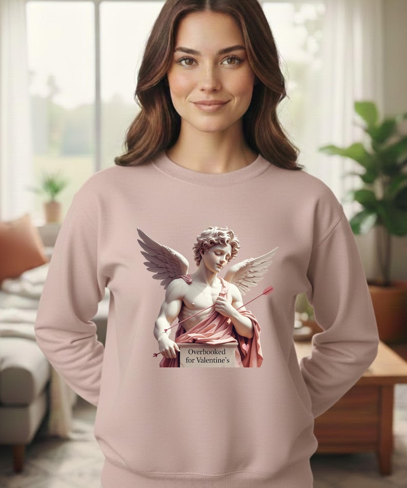 Puede incluir: Sudadera rosa claro con la imagen de un Cupido alado sosteniendo una flecha y un cartel con la frase "Overbooked for Valentine's". La sudadera es lisa y de manga larga.