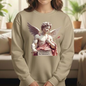 Puede incluir: Una sudadera beige con un gr&aacute;fico de una estatua cl&aacute;sica de Cupido, con alas y arco y flecha. La estatua est&aacute; cubierta con tela rosa y est&aacute; sobre un pedestal que dice "Overbooked for Valentine's".