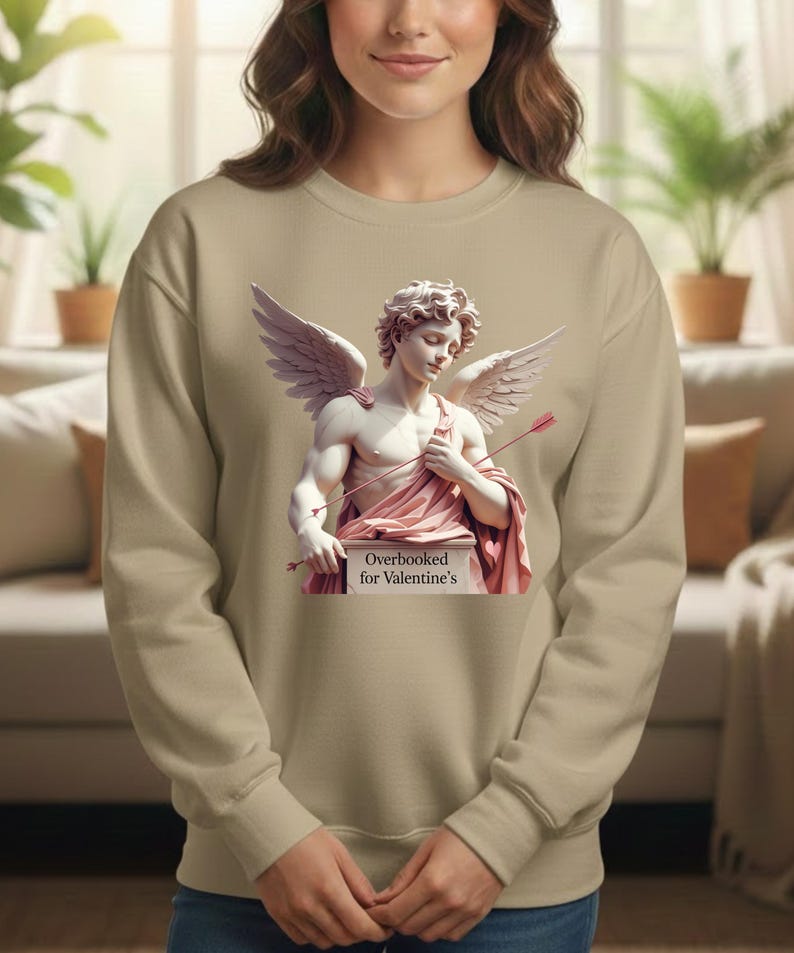 Puede incluir: Una sudadera beige con un gr&aacute;fico de una estatua cl&aacute;sica de Cupido, con alas y arco y flecha. La estatua est&aacute; cubierta con tela rosa y est&aacute; sobre un pedestal que dice "Overbooked for Valentine's".