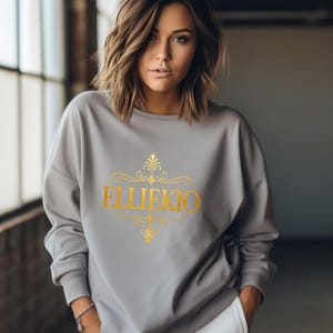 May include: A light gray long-sleeve sweatshirt featuring a gold-colored graphic print on the chest. The design includes the text ELLIEKIO in a bold serif font, centered between two ornate, symmetrical gold filigree flourishes above and below.