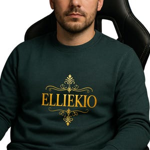 May include: A dark forest green crewneck sweatshirt featuring the text ELLIEKIO printed in a bold gold font. The text is centered on the chest and is framed by decorative gold filigree flourishes above and below the lettering.