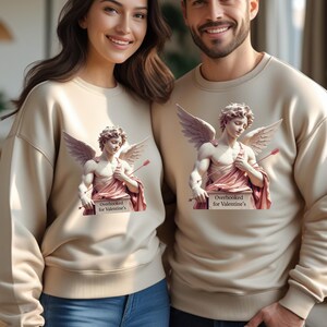 Puede incluir: Sudaderas beige con un dise&ntilde;o de estatua de Cupido y el texto "Overbooked for Valentine's". La estatua de Cupido tiene alas y sostiene una flecha. La pareja sonr&iacute;e y lleva vaqueros azules.