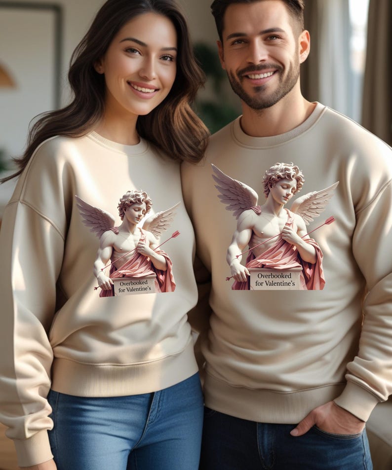 Puede incluir: Sudaderas beige con un dise&ntilde;o de estatua de Cupido y el texto "Overbooked for Valentine's". La estatua de Cupido tiene alas y sostiene una flecha. La pareja sonr&iacute;e y lleva vaqueros azules.