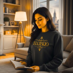 May include: A dark charcoal gray long-sleeve sweatshirt featuring the text ELLIEKIO in a bold, golden yellow serif font, accented by ornate, symmetrical golden scrollwork designs positioned directly above and below the centered lettering.