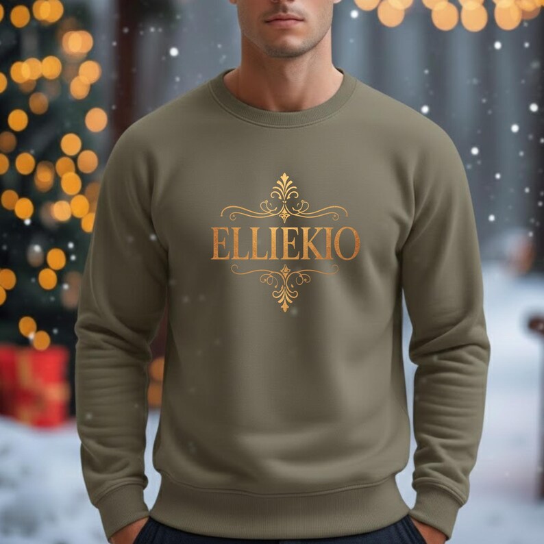 May include: A sage green crewneck sweatshirt featuring a gold-colored graphic print on the chest. The design includes the text ELLIEKIO in a serif font, centered between two ornate, symmetrical filigree flourishes above and below the lettering.