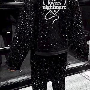 K&ouml;nnte beinhalten: Schwarzes Kapuzensweatshirt und passende Hose, beide mit funkelnden Verzierungen bedeckt. Auf der R&uuml;ckseite des Hoodies steht in Wei&szlig; der Text "lovers nightmare". Ein einzigartiges, auff&auml;lliges Ensemble.