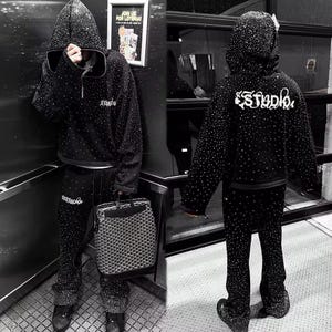 K&ouml;nnte beinhalten: Schwarzes Kapuzenpullover- und Hosen-Set, verziert mit zahlreichen kleinen, funkelnden Akzenten. Der Kapuzenpullover hat eine Kordelzugkapuze und das Wort "Studio" in wei&szlig;er Schrift auf dem R&uuml;cken. Die Hose hat ein ausgestelltes Bein-Design. Eine gemusterte Handtasche ist ebenfalls zu sehen.