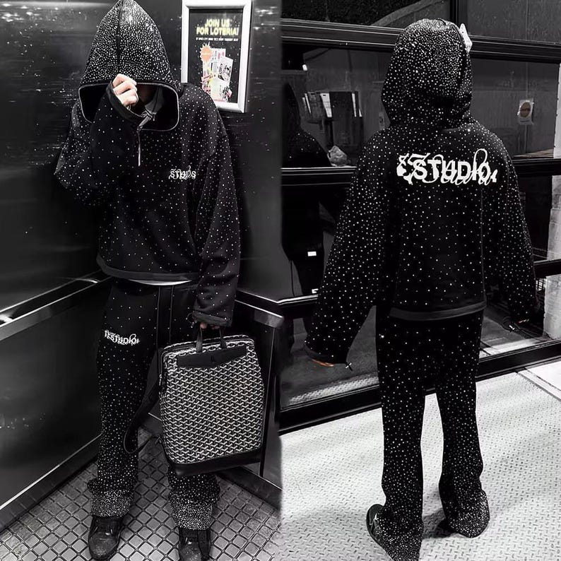 K&ouml;nnte beinhalten: Schwarzes Kapuzenpullover- und Hosen-Set, verziert mit zahlreichen kleinen, funkelnden Akzenten. Der Kapuzenpullover hat eine Kordelzugkapuze und das Wort "Studio" in wei&szlig;er Schrift auf dem R&uuml;cken. Die Hose hat ein ausgestelltes Bein-Design. Eine gemusterte Handtasche ist ebenfalls zu sehen.