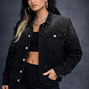 Puede incluir: Chaqueta vaquera negra adornada con pequeños tachuelas plateadas. La chaqueta tiene un cuello clásico, botones y bolsillos. Se muestra la marca "GOLDIVER". La modelo lleva un top y pantalones negros. Fondo gris oscuro.