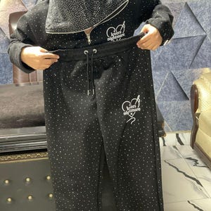 K&ouml;nnte beinhalten: Schwarzer Trainingsanzug mit Kapuzenjacke und passender Hose, beide mit zahlreichen kleinen, funkelnden Strasssteinen verziert. Die Jacke hat einen Rei&szlig;verschluss und den Text "lovers nightmare" in Wei&szlig;. Die Hose hat einen Kordelzug in der Taille.