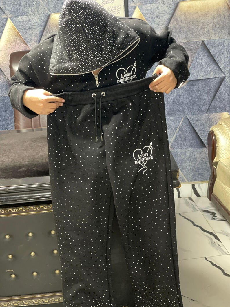K&ouml;nnte beinhalten: Schwarzer Trainingsanzug mit Kapuzenjacke und passender Hose, beide mit zahlreichen kleinen, funkelnden Strasssteinen verziert. Die Jacke hat einen Rei&szlig;verschluss und den Text "lovers nightmare" in Wei&szlig;. Die Hose hat einen Kordelzug in der Taille.