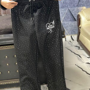K&ouml;nnte beinhalten: Schwarze Jogginghose mit wei&szlig;em Kordelzug und verstreuten Strasssteinen. Auf dem linken Bein steht in wei&szlig;er Schrift der Text "Lovers Nightmare". Eine passende Jacke ist im Hintergrund zu sehen.