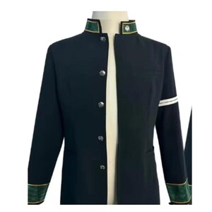 Puede incluir: Chaqueta negra con cuello mandarín, botones plateados y ribetes verdes y dorados en el cuello y los puños. La manga izquierda tiene una franja blanca. La chaqueta se exhibe en un maniquí.