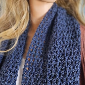 May include: A close-up of a navy blue crochet scarf. The scarf is made of a soft, textured yarn and has an open, lacy design. The scarf is draped around the neck and shoulders.