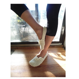 May include: A pair of white crocheted slippers with a pearl button detail. The slippers are worn on a pair of legs wearing black leggings.