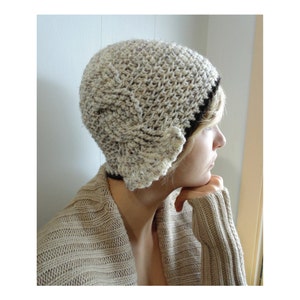 May include: A close-up of a woman wearing a beige crocheted hat with a black trim. The hat has a unique design with a folded flap on the side. The woman is wearing a beige sweater and has her hand on her chin.