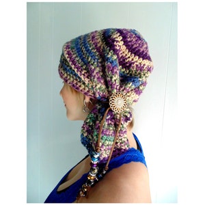 May include: A crocheted hat with a striped pattern in shades of purple, green, and beige. The hat is tied at the back with a brown cord and a decorative metal flower pin.