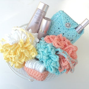 May include: A white wire basket filled with crocheted bath items. There are three crocheted flower-shaped bath poufs in yellow, white, and peach. There is a small crocheted bag with a silver button and two bottles of lotion.
