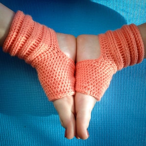 May include: A pair of coral-colored crocheted ankle warmers with a hole for the heel. The warmers are worn on a person's feet and are shown against a blue background.