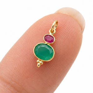 May include: A gold pendant featuring a small oval-shaped pink gemstone positioned above a larger oval-shaped green gemstone. The pendant has a small gold loop at the top for attachment and a decorative gold bead detail at the bottom base.