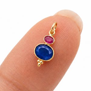 Puede incluir: Un colgante de oro con una gran piedra preciosa azul de talla ovalada y una piedra preciosa rosa más pequeña de talla redonda. El colgante tiene una pequeña anilla de oro en la parte superior y un detalle de cuenta de oro bajo la piedra azul.