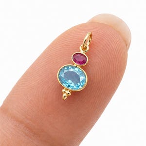 Puede incluir: Un colgante de oro con una gema azul grande de talla ovalada y una gema rosa más pequeña de talla redonda dispuestas verticalmente. El colgante incluye una pequeña anilla de oro en la parte superior y una cuenta decorativa de oro en la base.