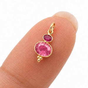 May include: A small gold pendant featuring two oval-shaped pink gemstones set in a vertical arrangement. The larger stone is at the bottom, and the smaller stone is at the top, connected by a gold jump ring for attachment to a chain or necklace.