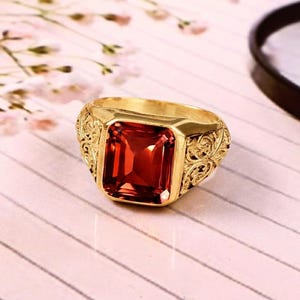 Könnte beinhalten: Ein goldener Ring mit einem großen, rechteckigen, facettierten roten Edelstein in der Mitte. Der Ring besteht aus Gold und weist auf beiden Seiten der zentralen Steinfassung komplizierte, erhabene Schnörkelmuster auf.