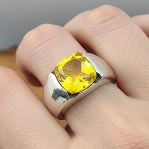 Può includere: Un anello d'argento con una grande gemma gialla brillante a taglio quadrato. L'anello ha una fascia ampia e lucida e un design moderno. La gemma è il punto focale, catturando la luce e aggiungendo un tocco di colore.