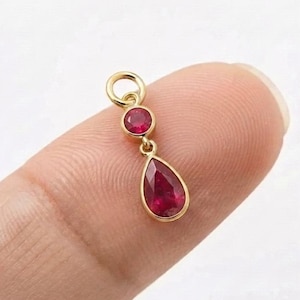 Pode incluir: Um pingente de ouro com um pequeno rubi redondo engastado sobre uma gota de rubi em forma de pera. O pingente inclui um anel circular no topo para fixação, exibindo duas pedras preciosas de cor vermelha vibrante em ouro amarelo polido.